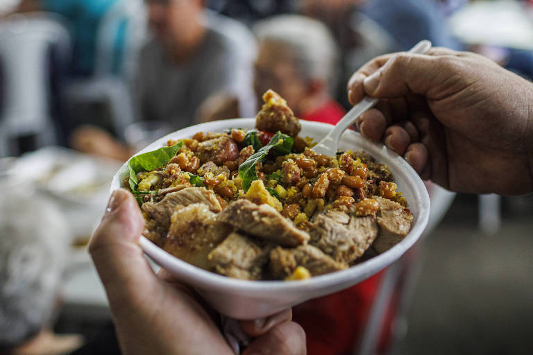 A imagem mostra uma mão segurando um prato de comida， que contém arroz， carne e vegetais. Ao fundo， há uma grande quantidade de pessoas sentadas em mesas， em um ambiente movimentado， possivelmente um evento ou feira de comida.