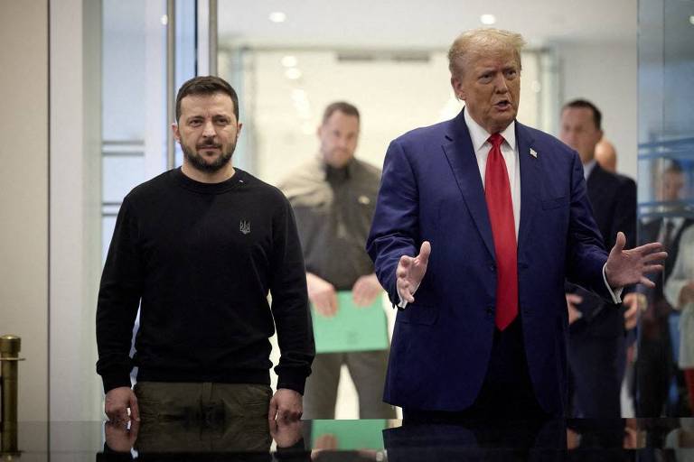 A imagem mostra dois homens em um ambiente interno. À esquerda， um homem com cabelo curto e barba， vestindo uma camiseta preta com o símbolo da Ucrânia， está em pé ao lado de um homem mais alto， com cabelo loiro e terno azul， que gesticula com as mãos. Ao fundo， há outras pessoas visíveis， mas não claramente identificáveis.