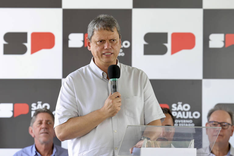 Tarcísio de Freitas， homem de camisa branca， está falando ao microfone em um evento. Ele parece sério e está em frente a um púlpito de vidro. Ao fundo， há um painel com as letras 039;SP039; em preto e vermelho， e o nome 039;SÃO PAULO039; em destaque. Dois homens estão sentados atrás dele， um deles olhando para frente e o outro com uma expressão pensativa