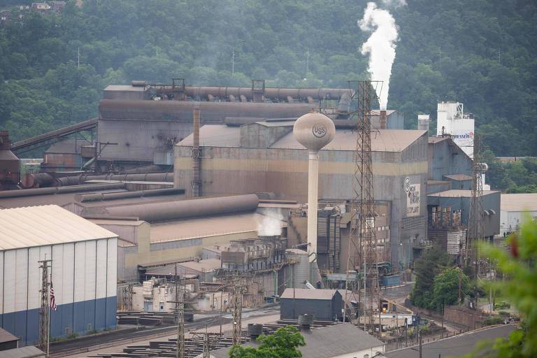 A imagem mostra uma grande instalação industrial com várias estruturas metálicas e um grande tanque de armazenamento. Ao fundo， há uma montanha coberta de vegetação. Uma figura branca， possivelmente uma estátua ou uma chaminé， se destaca no topo da fábrica， emitindo vapor. O céu está nublado， e a cena transmite uma atmosfera de atividade industrial.