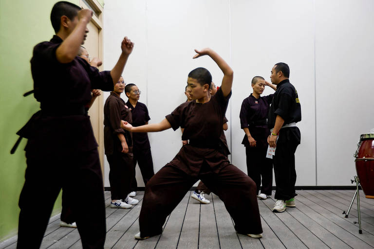 Um grupo de pessoas em um ambiente interno， praticando movimentos de artes marciais. Algumas estão em posições de combate， enquanto outras observam. Todos estão vestidos com roupas escuras， e o chão é de madeira. Ao fundo， há um instrumento musical de percussão.
