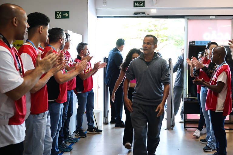 A imagem mostra um grupo de pessoas aplaudindo enquanto um homem caminha em direção à saída de um local. As pessoas estão vestidas com camisetas vermelhas e algumas usam calças jeans. O homem está vestido com uma camiseta cinza e calças escuras. Ao fundo, há uma porta de saída e uma máquina de vendas. A atmosfera é de celebração e acolhimento.