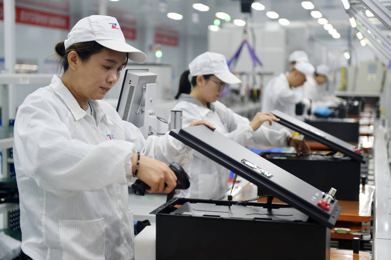 A imagem mostra várias mulheres trabalhando em uma linha de montagem em uma fábrica. Elas estão vestindo uniformes brancos e bonés， e estão concentradas em suas tarefas， manipulando equipamentos e componentes eletrônicos. O ambiente é bem iluminado e organizado， com mesas de trabalho e ferramentas visíveis ao fundo.