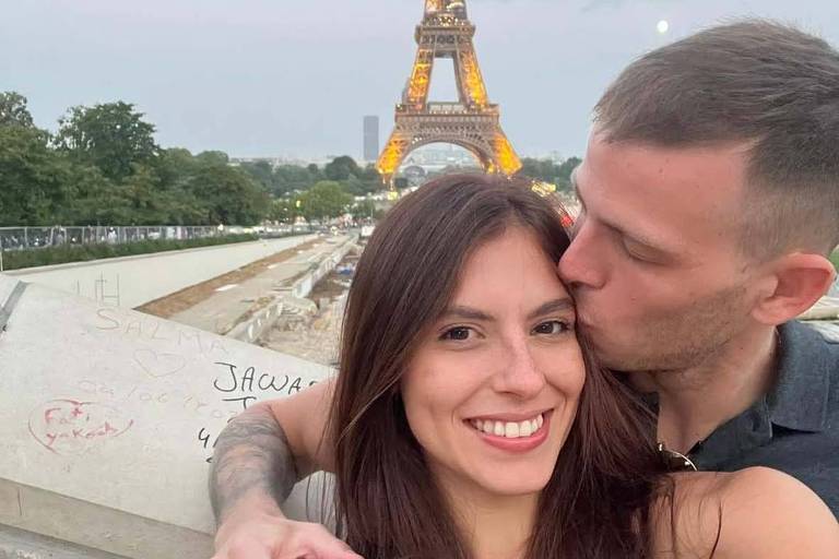 Foto colorida mostra um homem e uma mulher caucasianos. Ele tem cabelo curto e ela cabelos longos. O homem está dando um beijo no rosto da mulher. Ao fundo aparece a Torre Eifel