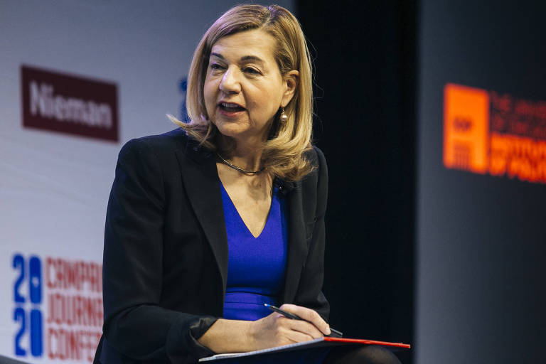 Uma mulher com cabelo loiro e liso， vestindo uma blusa azul e um blazer preto， está sentada em um palco durante uma conferência. Ela parece estar falando ou respondendo a uma pergunta， segurando uma caneta e um bloco de notas. Ao fundo， há elementos gráficos e logotipos relacionados ao evento.