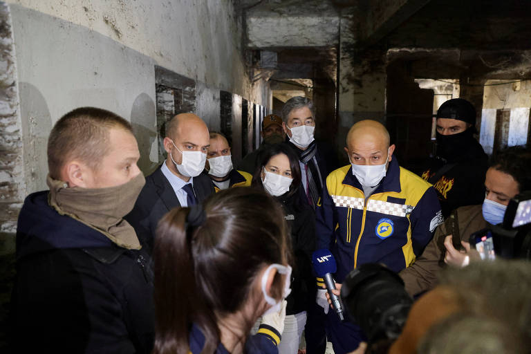 Um grupo de pessoas， algumas usando máscaras， está reunido em um ambiente interno com paredes desgastadas e um aspecto industrial. Algumas pessoas estão de frente， enquanto outras estão parcialmente visíveis. O grupo parece estar em uma conversa ou reunião， com algumas pessoas segurando dispositivos móveis.
