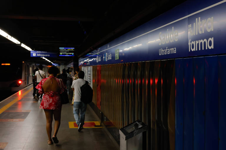 A imagem mostra uma plataforma de metrô com pessoas caminhando. À esquerda， uma mulher com um vestido rosa e， à direita， um homem com uma mochila. O fundo apresenta uma parede azul com o nome da estação visível. Há também iluminação artificial e um lixeiro na plataforma