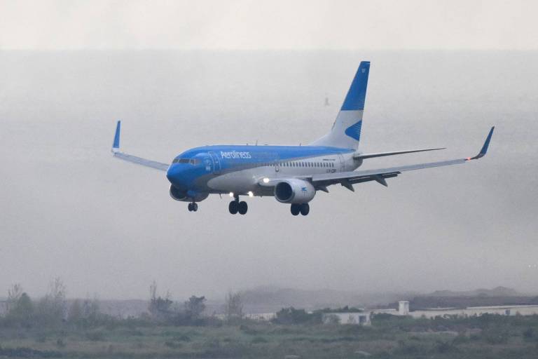 Um avião comercial， com fuselagem azul e branca， está em processo de aterrissagem. O céu está nublado， e a visibilidade parece limitada. O avião está inclinado levemente para baixo， com as rodas visíveis， prestes a tocar o solo.