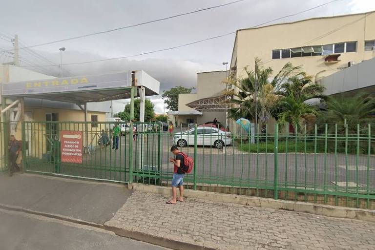 Imagem mostra a fachada do hospital， com grades em verde， pessoas passando na frente e carros estacionados