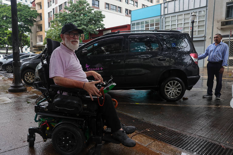 Um homem idoso com barba branca e óculos está sentado em uma cadeira de rodas motorizada. Ele usa uma camisa rosa e um boné preto. Ao fundo, há um carro preto estacionado e um homem em pé ao lado do veículo, aparentemente ajudando. O ambiente é urbano, com prédios e árvores ao fundo, e o chão está molhado, sugerindo que pode ter chovido.