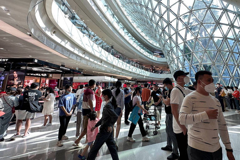 A imagem mostra o interior de um shopping moderno， com um design arquitetônico arrojado. O teto é composto por uma estrutura de vidro e metal， permitindo a entrada de luz natural. Há várias pessoas caminhando pelo espaço， algumas paradas em lojas. O piso é de cerâmica brilhante， refletindo a luz. O ambiente é amplo e arejado， com várias áreas de compras visíveis ao fundo.