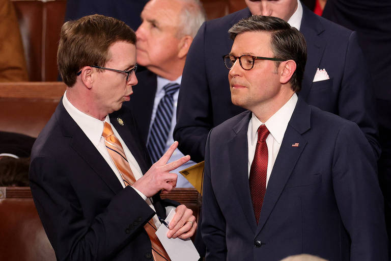 A imagem mostra dois homens em um ambiente legislativo， aparentemente conversando. Um deles está usando um terno escuro e uma gravata vermelha， enquanto o outro veste um terno escuro com uma gravata laranja. O homem com a gravata laranja está gesticulando com a mão， enquanto o outro observa atentamente. Ao fundo， é possível ver outras pessoas e o ambiente típico de uma câmara legislativa.