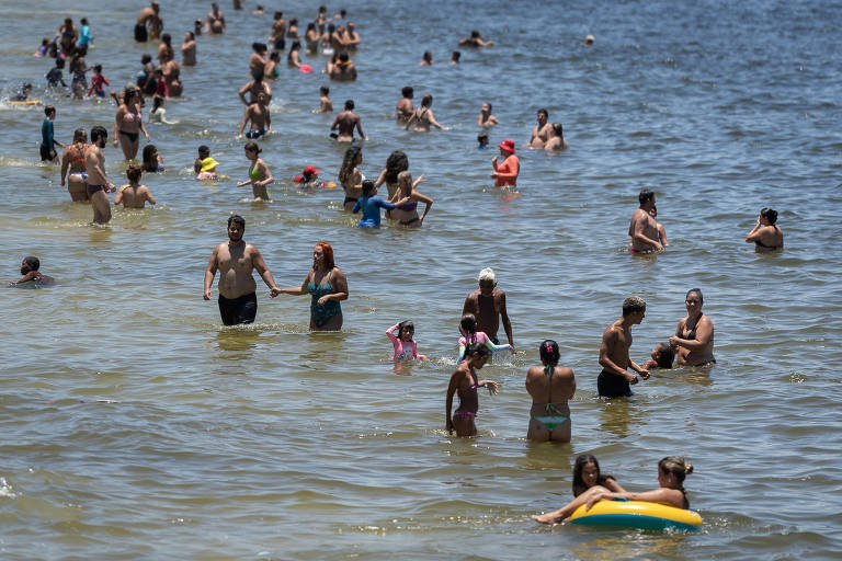 A imagem mostra uma praia lotada de pessoas se divertindo na água. Há adultos e crianças, alguns estão nadando, enquanto outros estão em pé na água. A cena é ensolarada, com várias pessoas usando roupas de banho. Algumas crianças estão brincando com boias e outras estão sendo seguradas por adultos.