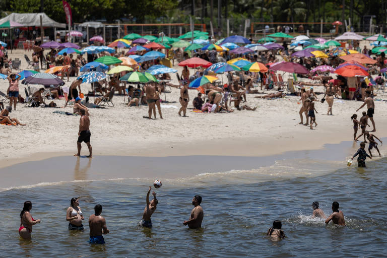 A imagem mostra uma praia cheia de pessoas. Há uma grande quantidade de guarda-sóis coloridos， predominantemente em tons de verde， amarelo， vermelho e azul， cobrindo a areia. Algumas pessoas estão na água， enquanto outras estão na areia， relaxando ou socializando. O ambiente é ensolarado e vibrante， com uma atmosfera de lazer e diversão.
