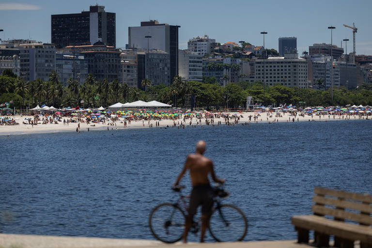 A imagem mostra um ciclista em primeiro plano， parado ao lado de sua bicicleta， com o mar ao fundo. Ao longe， é possível ver uma praia cheia de pessoas e a cidade com prédios altos e vegetação. O céu está claro e ensolarado.