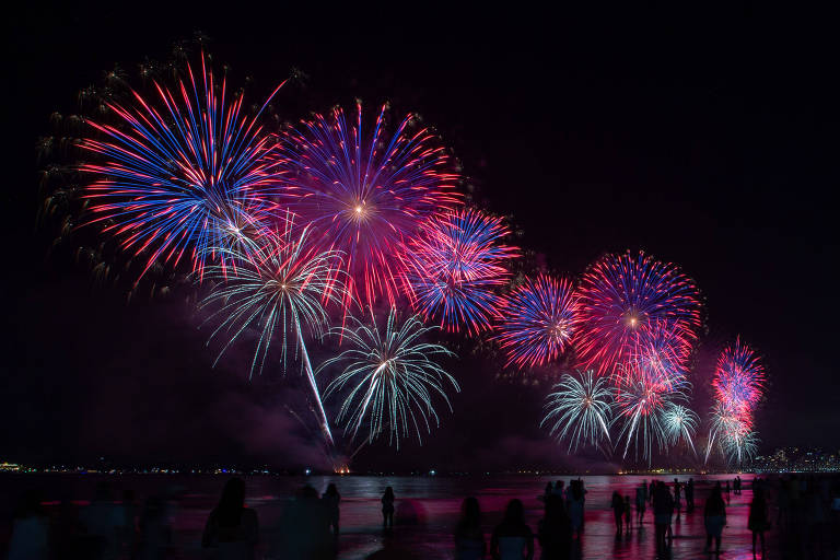 A imagem mostra um espetáculo de fogos de artifício iluminando o céu noturno. Os fogos são predominantemente nas cores vermelha， azul e branca， criando um efeito visual vibrante. Na parte inferior da imagem， há silhuetas de pessoas observando o show， em uma praia com reflexos na água