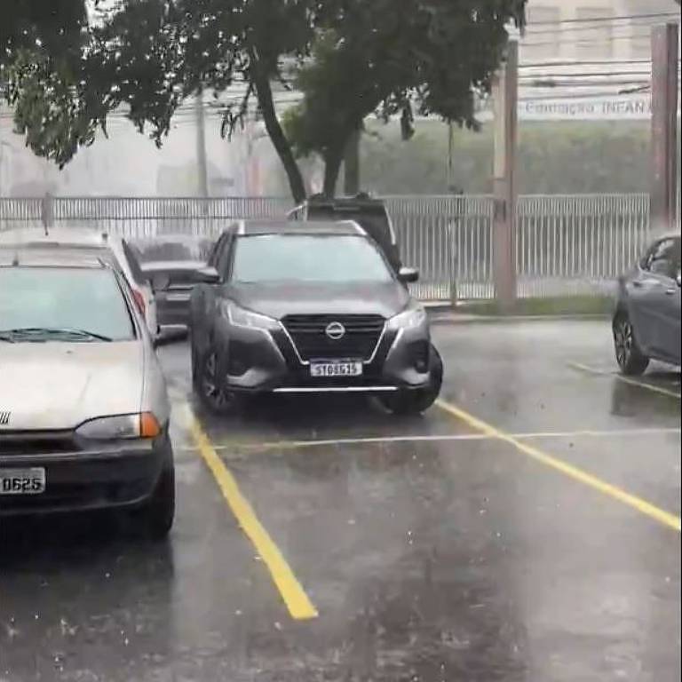 A imagem mostra um estacionamento durante uma forte chuva. Um carro preto está estacionado em uma das vagas， enquanto outros carros estão visíveis ao fundo. A chuva é intensa， e a água está acumulada no chão， refletindo a luz. Árvores estão ao redor， e o ambiente parece nublado.