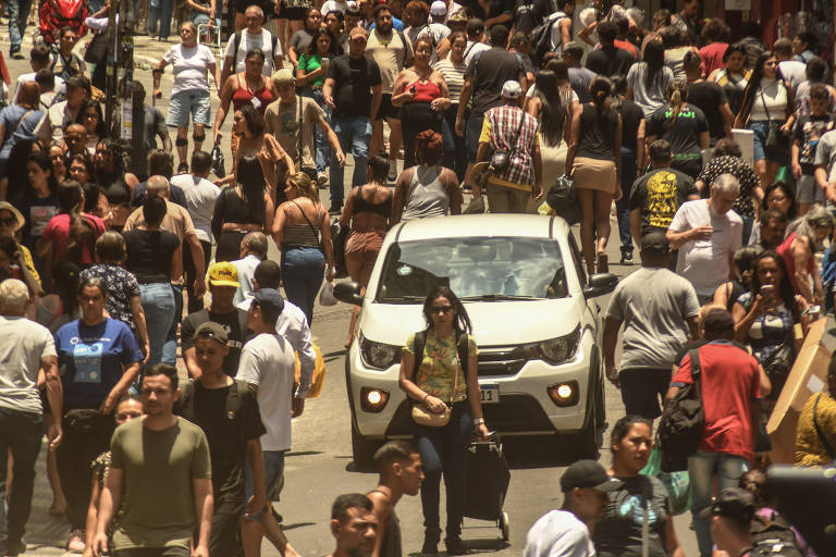 Imagem mostra uma multidão andando por uma rua， lotando tanto as calçadas quanto a via. Um carro branco tenta passar， sendo cercado pelos transeuntes