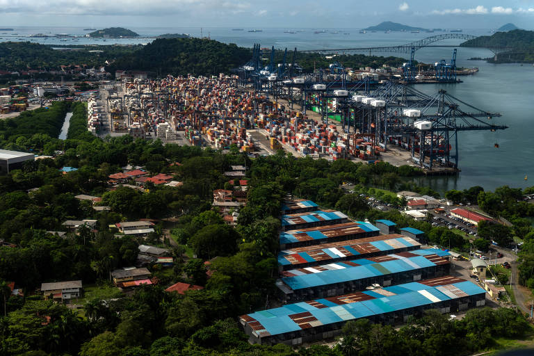 A imagem mostra uma vista aérea de um porto movimentado， com contêineres coloridos empilhados em um terminal. À esquerda， há uma área urbana com casas e vegetação densa. No fundo， é possível ver um rio que flui em direção ao mar， além de uma ponte que conecta as duas margens. O céu está parcialmente nublado e há navios ancorados no mar.