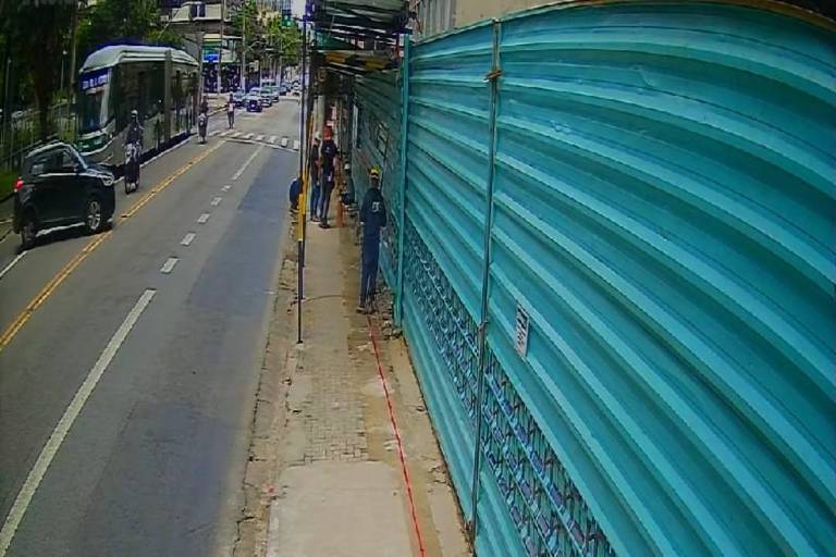 A imagem mostra uma rua urbana com um canteiro de obras à direita. Há um muro de proteção azul ao longo da calçada， onde algumas pessoas estão trabalhando. Um ônibus e um carro preto estão passando pela rua， enquanto motociclistas também estão visíveis