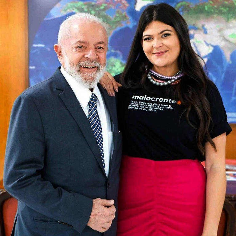 Na imagem， duas pessoas estão posando para uma foto em um ambiente interno. À esquerda， um homem idoso com cabelo grisalho e barba， vestindo um terno escuro e uma gravata listrada. À direita， uma mulher com cabelo longo e escuro， usando uma blusa preta com a palavra 039;malocren039;te039; e uma saia rosa. Ao fundo， há um mapa-múndi e um texto que diz 039;ÁREAS PROTEGIDAS NO MUNDO039;.