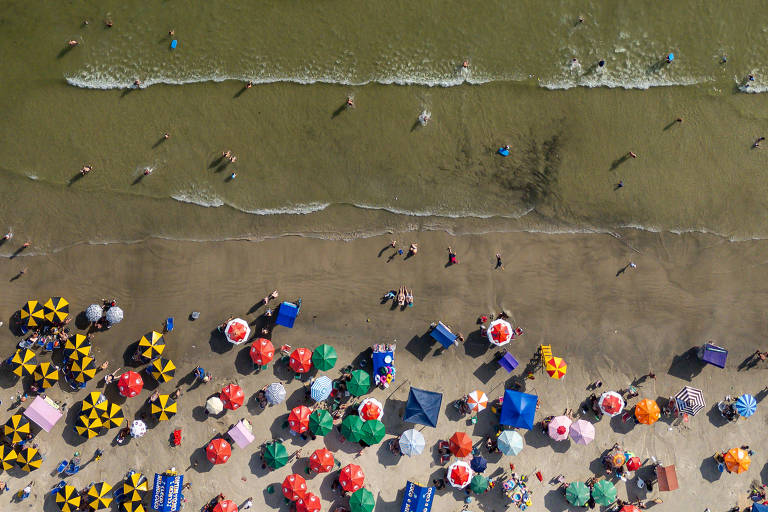 A imagem mostra uma vista aérea de uma praia， onde várias pessoas estão deitadas em toalhas e sob guarda-sóis coloridos. O chão da praia é de areia clara， e há uma linha de água visível ao fundo. A área ao redor dos guarda-sóis é preenchida com toalhas em diversas cores， criando um padrão vibrante.
