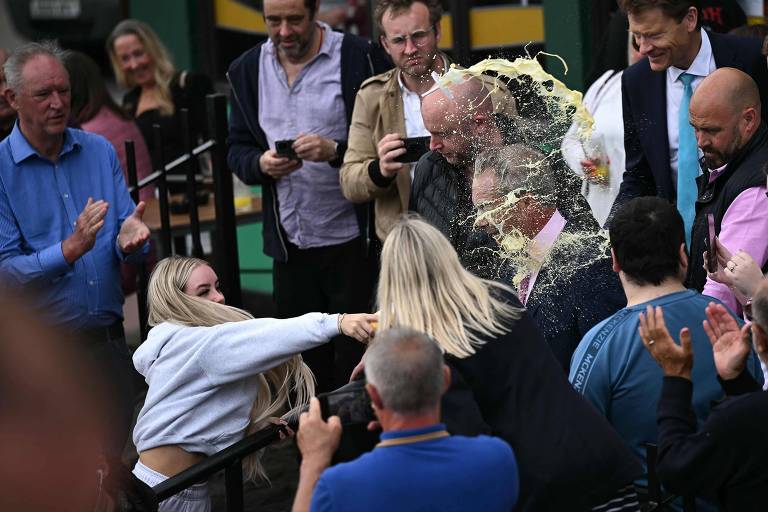 A imagem mostra um grupo de pessoas em um evento， onde uma mulher de cabelo loiro está jogando um líquido sobre um homem. O homem está cercado por outras pessoas que observam a cena， algumas aplaudindo. O ambiente parece ser festivo， com várias pessoas usando roupas casuais e sorrindo. Ao fundo， há uma multidão assistindo à cena.