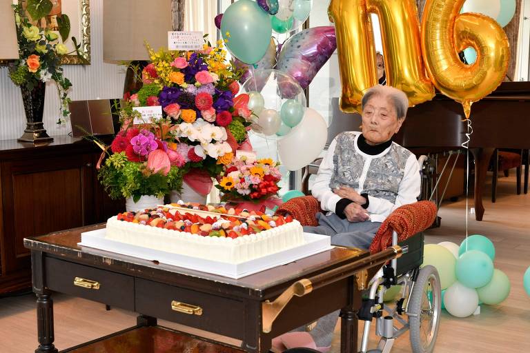 A imagem mostra uma mulher idosa sentada em uma cadeira de rodas， comemorando seu 116º aniversário. Ela está ao lado de um grande bolo de aniversário decorado com frutas e creme. Ao fundo， há um arranjo de flores coloridas e balões em tons de verde e branco. Os números 039;116039; em balões dourados estão destacados na imagem.