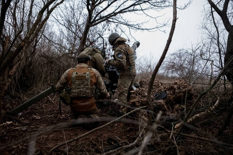 A imagem mostra um grupo de soldados em um ambiente florestal， com árvores secas ao fundo. Dois soldados estão agachados， enquanto um terceiro está em pé， aparentemente ajustando um equipamento. Todos estão vestidos com uniformes militares camuflados e capacetes. O solo é coberto por folhas secas e galhos， criando um cenário de camuflagem.