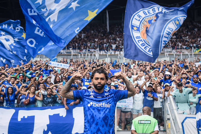 Um jogador de futebol está em destaque no primeiro plano， fazendo pose com os braços flexionados. Ele usa uma camisa azul com detalhes brancos e o logo da Betfair. Ao fundo， uma grande torcida está reunida， segurando bandeiras azuis e brancas， com muitos torcedores vestindo camisas do time. O ambiente é de celebração e apoio ao time.