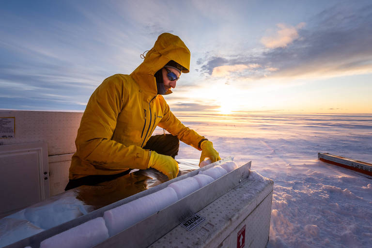 Um homem vestido com um casaco amarelo e luvas， trabalhando em um ambiente coberto de neve. Ele está manipulando um equipamento que parece ser uma caixa de gelo， enquanto o sol se põe ao fundo， iluminando o céu com tons de azul e laranja. O cenário é vasto， com neve se estendendo até onde a vista alcança.