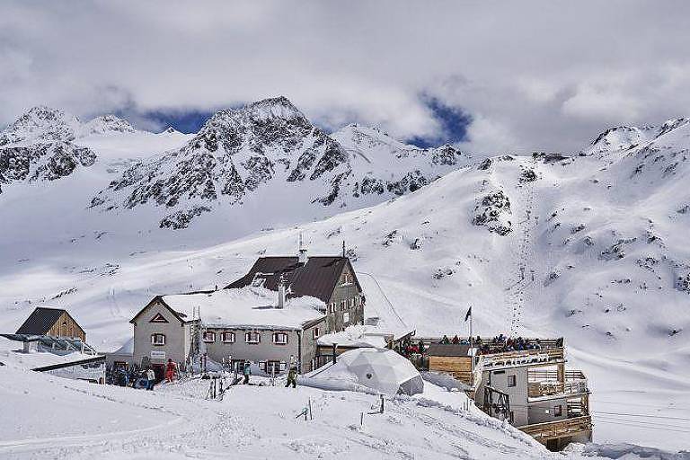 A imagem mostra uma paisagem montanhosa coberta de neve， com várias montanhas ao fundo sob um céu nublado. No primeiro plano， há um edifício de dois andares， que parece ser uma cabana ou refúgio de montanha， cercado por neve. 