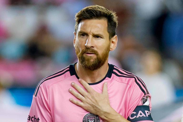 Um jogador de futebol está em campo， vestindo uma camisa rosa com detalhes pretos. Ele tem uma barba e cabelo curto， e está fazendo um gesto com a mão sobre o peito， parecendo pensativo. Ao fundo， há uma multidão desfocada.