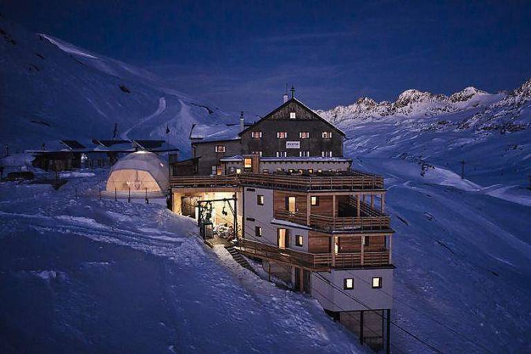 O albergue quot;Linda vista - Bella Vistaquot;. A imagem mostra um chalé de madeira e pedra iluminado à noite， cercado por neve e montanhas ao fundo.