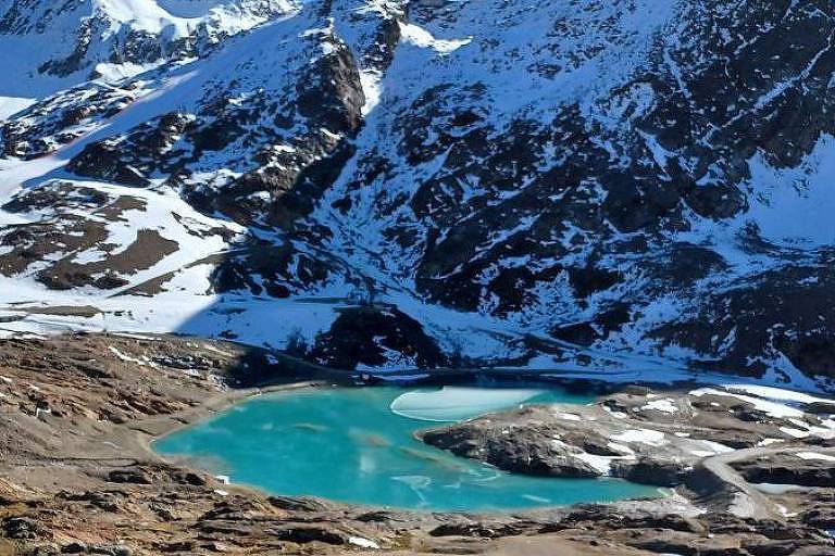 A imagem mostra a geleira Hochjochferner no outono， parcialmente derretida， com um lago turquesa ao centro， cercada por montanhas cobertas de neve e terreno rochoso exposto.