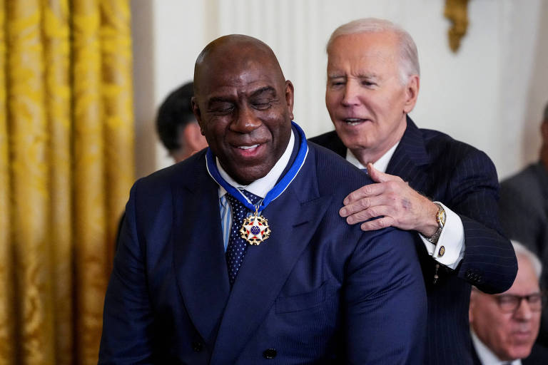 A imagem mostra um homem sorridente recebendo uma medalha de honra， enquanto um outro homem， que está atrás dele， coloca a mão em seu ombro. O homem à frente usa um terno azul e uma gravata com bolinhas， e a medalha pendurada em seu pescoço tem um design elaborado. Ao fundo， há cortinas amarelas e outras pessoas assistindo à cerimônia.