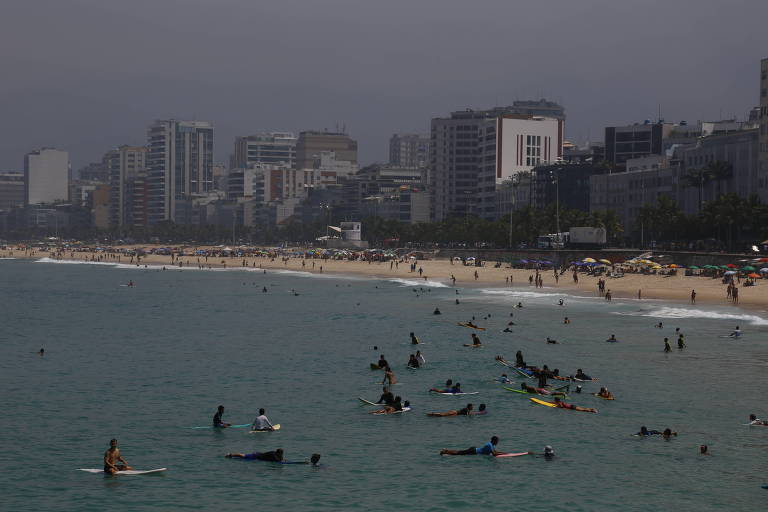 A imagem mostra uma praia com várias pessoas surfando e nadando no mar. Ao fundo， há edifícios altos e uma faixa de areia onde algumas pessoas estão presentes. A água é de um tom azul esverdeado.