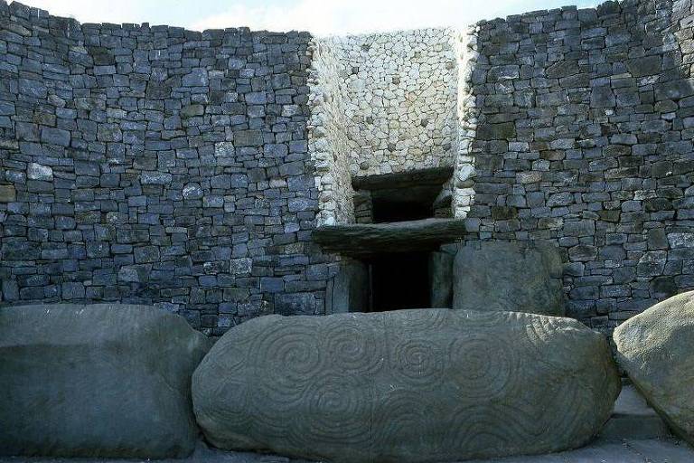 A imagem mostra uma estrutura de pedra antiga， com paredes de pedras empilhadas formando um espaço interno. No centro， há uma abertura que parece levar a um espaço mais profundo， e várias pedras grandes estão dispostas ao redor da entrada. A luz natural entra pela abertura superior， iluminando o interior da construção.