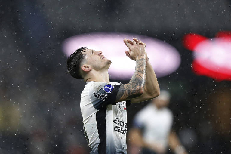 Um jogador de futebol está de pé， com as mãos unidas em oração， olhando para cima. Ele está molhado devido à chuva， e seu uniforme é predominantemente branco com detalhes em preto. O fundo é desfocado， com luzes e gotas de água visíveis no ar， sugerindo um ambiente de jogo intenso.