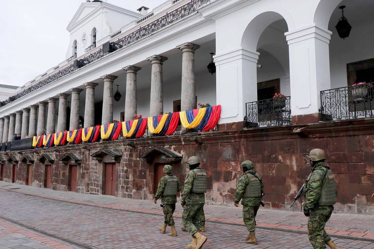 A imagem mostra soldados marchando em frente a um edifício histórico com colunas. O edifício possui bandeiras coloridas penduradas em sua fachada， predominantemente nas cores da bandeira do Equador. O céu está nublado， e a cena transmite um ambiente solene.