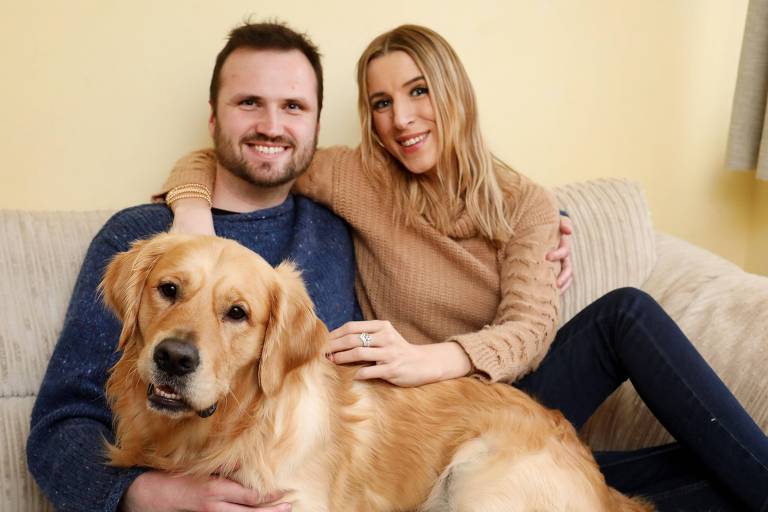 Um homem e uma mulher estão sentados em um sofá. O homem está usando um suéter azul e a mulher está vestindo um suéter bege. Eles estão sorrindo e o homem está segurando um cachorro da raça Golden Retriever， que está sentado em seu colo. O ambiente é iluminado e as paredes são de uma cor clara.