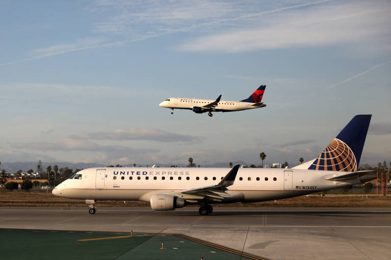 A imagem mostra dois aviões em um aeroporto. Um avião da United Airlines está taxiando na pista， enquanto um avião da Delta Airlines está sobrevoando， prestes a pousar. O céu está parcialmente nublado e há árvores ao fundo.