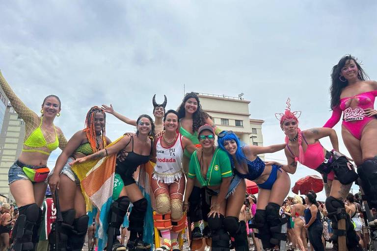 Um grupo de mulheres posando para a foto durante uma festa de Carnaval. Elas estão vestidas com trajes coloridos e variados， incluindo biquínis e acessórios festivos. Algumas usam patins e outras têm cabelos de cores diferentes. O fundo mostra um ambiente de festa com prédios e guarda-sóis.