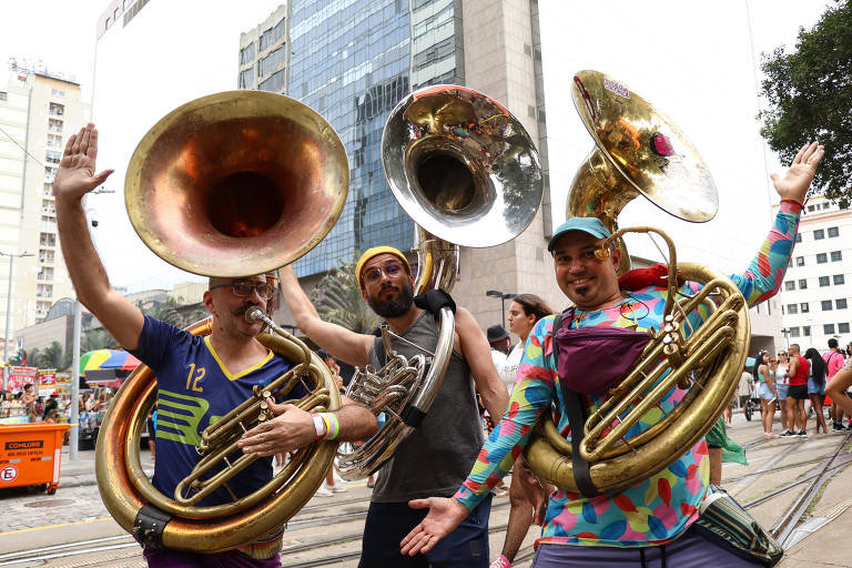 Três músicos estão posando para a foto durante um evento de Carnaval. Cada um deles segura uma tuba. O primeiro músico à esquerda usa uma camiseta colorida com o número 12 e tem um braço levantado. O segundo， ao centro， está sem camisa e usa óculos escuros. O terceiro， à direita， veste uma camiseta estampada e também levanta um braço. Ao fundo， há prédios altos e uma multidão participando do evento.