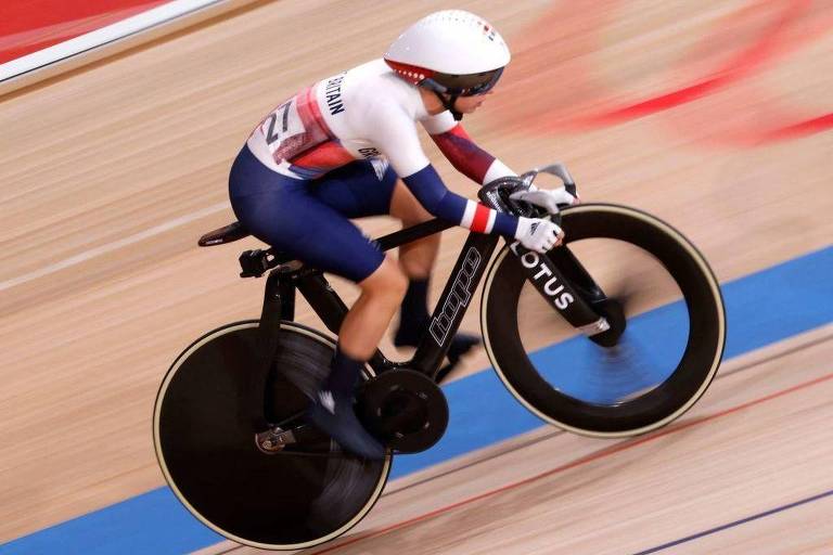 Uma ciclista está competindo em uma pista de ciclismo, usando um capacete branco e um uniforme com as cores da bandeira nacional. A bicicleta tem rodas grandes e pretas, e o ciclista está em uma posição aerodinâmica, pedalando rapidamente. O fundo mostra a pista de madeira e as marcas de corrida.