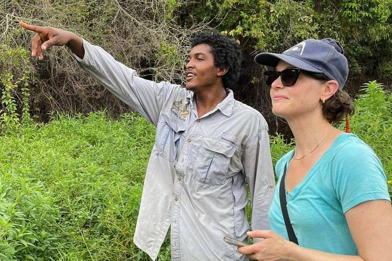 A imagem mostra um homem e uma mulher em um ambiente natural. O homem， com pele escura e cabelo encaracolado， está apontando para algo à sua frente， enquanto a mulher， com cabelo curto e usando um boné， observa atentamente. Ambos estão cercados por vegetação densa e verde.