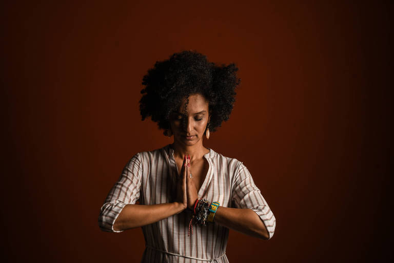 A imagem mostra uma mulher com cabelo afro， vestindo uma blusa de listras， em uma pose de meditação com as mãos unidas em frente ao peito. O fundo é de uma cor marrom escura， criando um contraste com a figura da mulher.