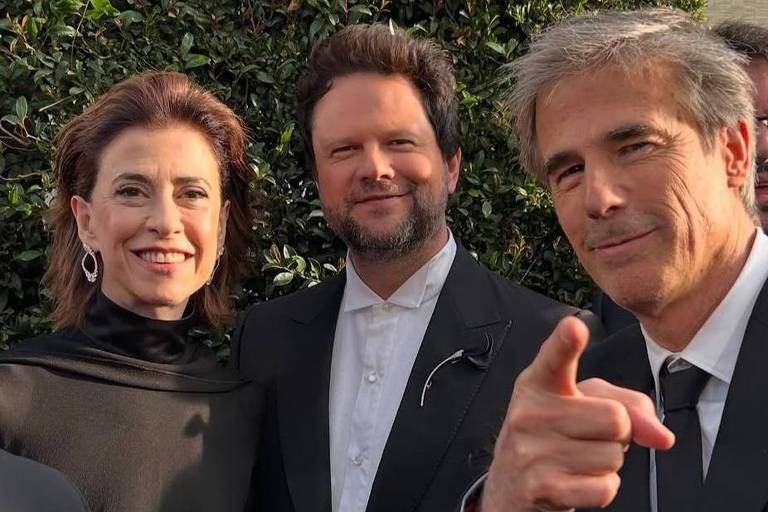 Três pessoas posam para a foto em um evento, com um fundo de folhagem verde. A mulher à esquerda usa um vestido preto e brincos grandes, sorrindo. O homem ao centro, com cabelo cacheado e barba, veste um terno preto e uma camisa branca. O homem à direita, com cabelo grisalho, aponta para a câmera e também está vestido de terno preto. Ao fundo, é possível ver outras pessoas e uma estrutura arquitetônica.