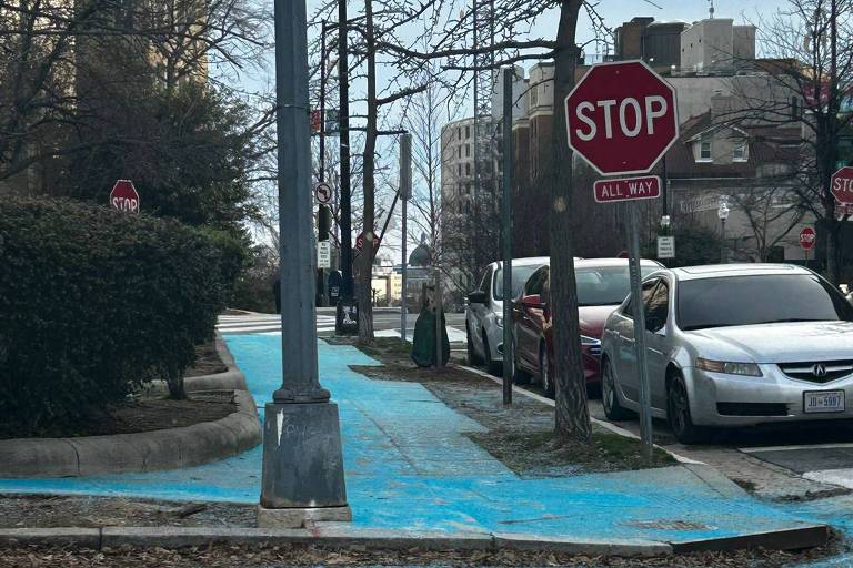 A imagem mostra uma calçada pintada de azul， que é sal， com um sinal de 039;STOP039; visível ao fundo. Há postes de luz e árvores ao lado da calçada， e alguns veículos estacionados na rua. O céu está nublado.