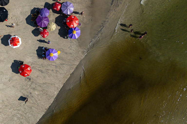 Imagem aérea mostra um afluente de um córrego com água turva desaguando no mar. Ao lado dá afluente há alguns guarda-sóis laranjas e roxos. 